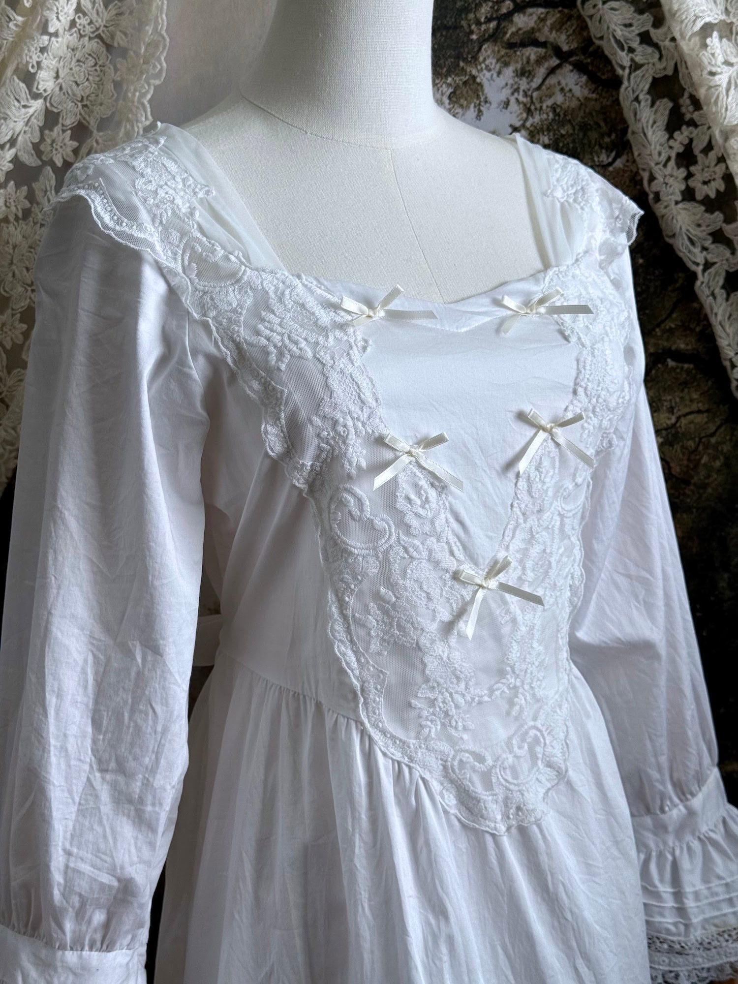A historically-inspired 1700s style white cotton basque waist night dress with bell sleeves and lace trim pictured on a mannequin in front of an art-history backdrop.