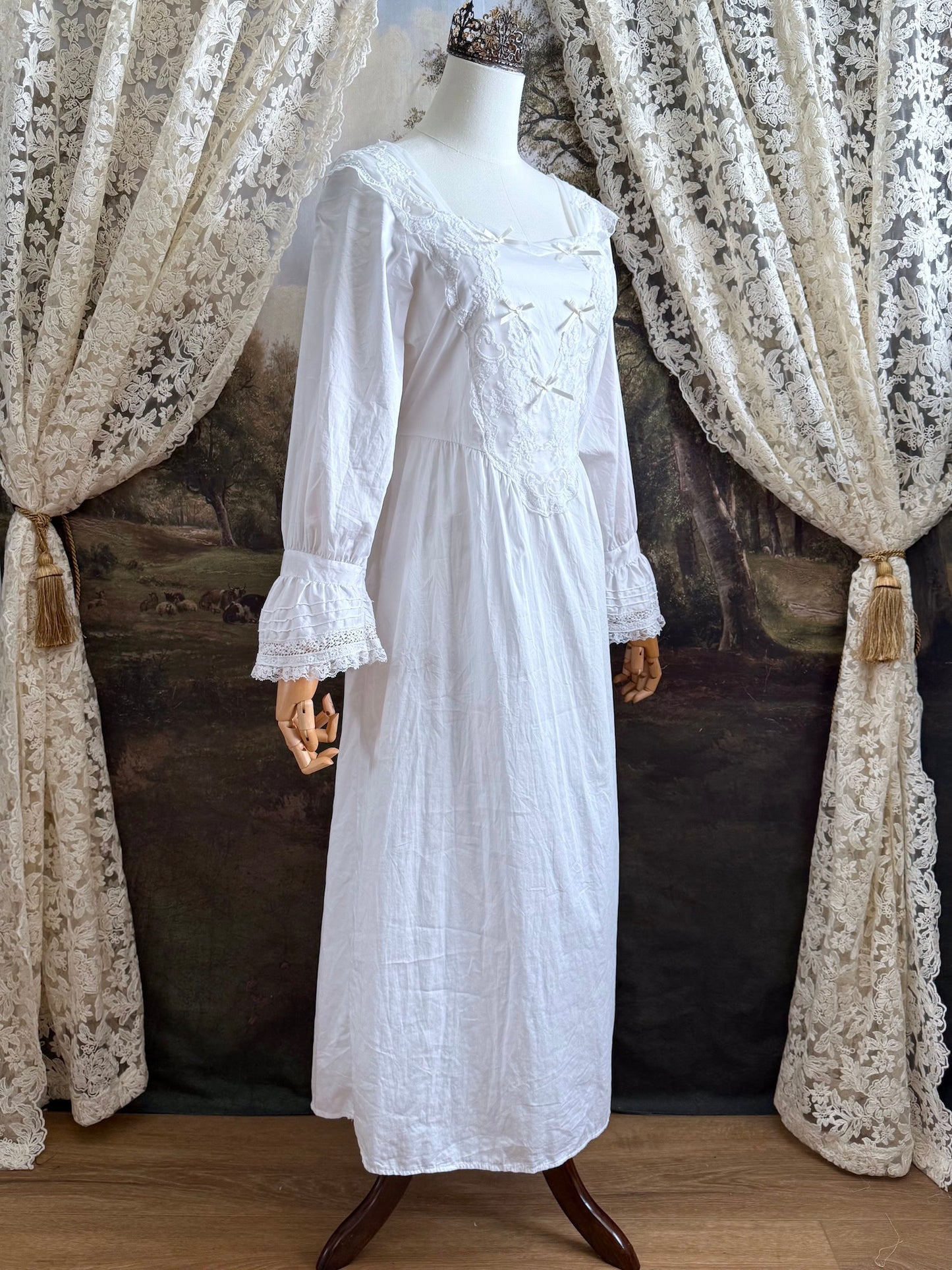 A historically-inspired 1700s style white cotton basque waist night dress with bell sleeves and lace trim pictured on a mannequin in front of an art-history backdrop.