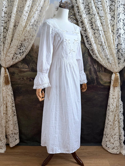 A historically-inspired 1700s style white cotton basque waist night dress with bell sleeves and lace trim pictured on a mannequin in front of an art-history backdrop.
