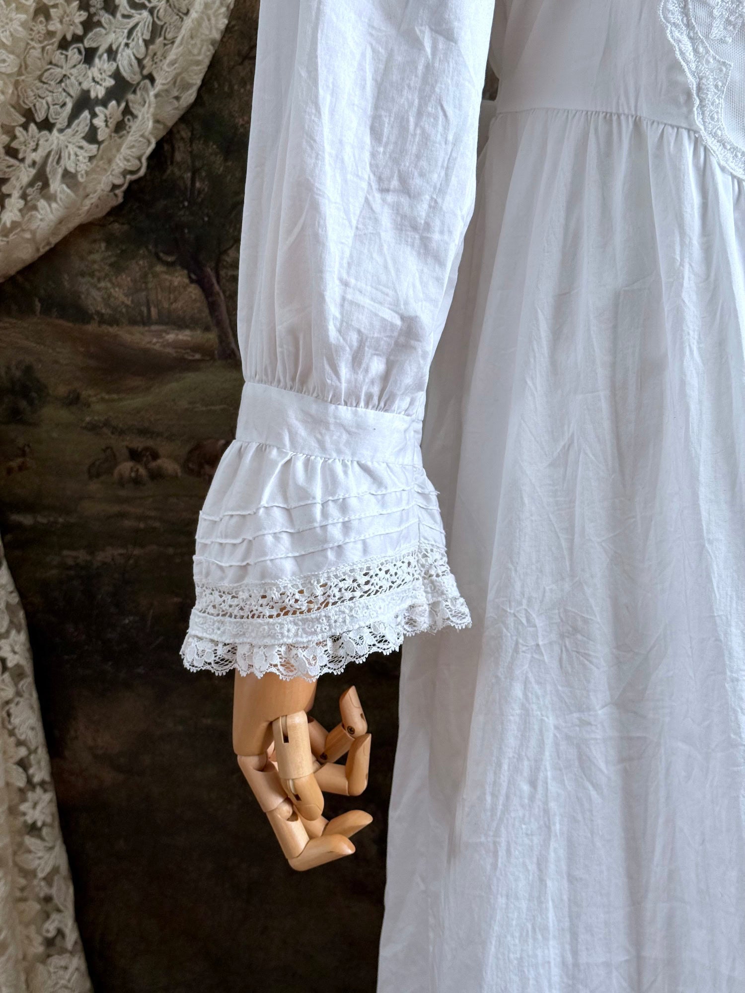 A historically-inspired 1700s style white cotton basque waist night dress with bell sleeves and lace trim pictured on a mannequin in front of an art-history backdrop.