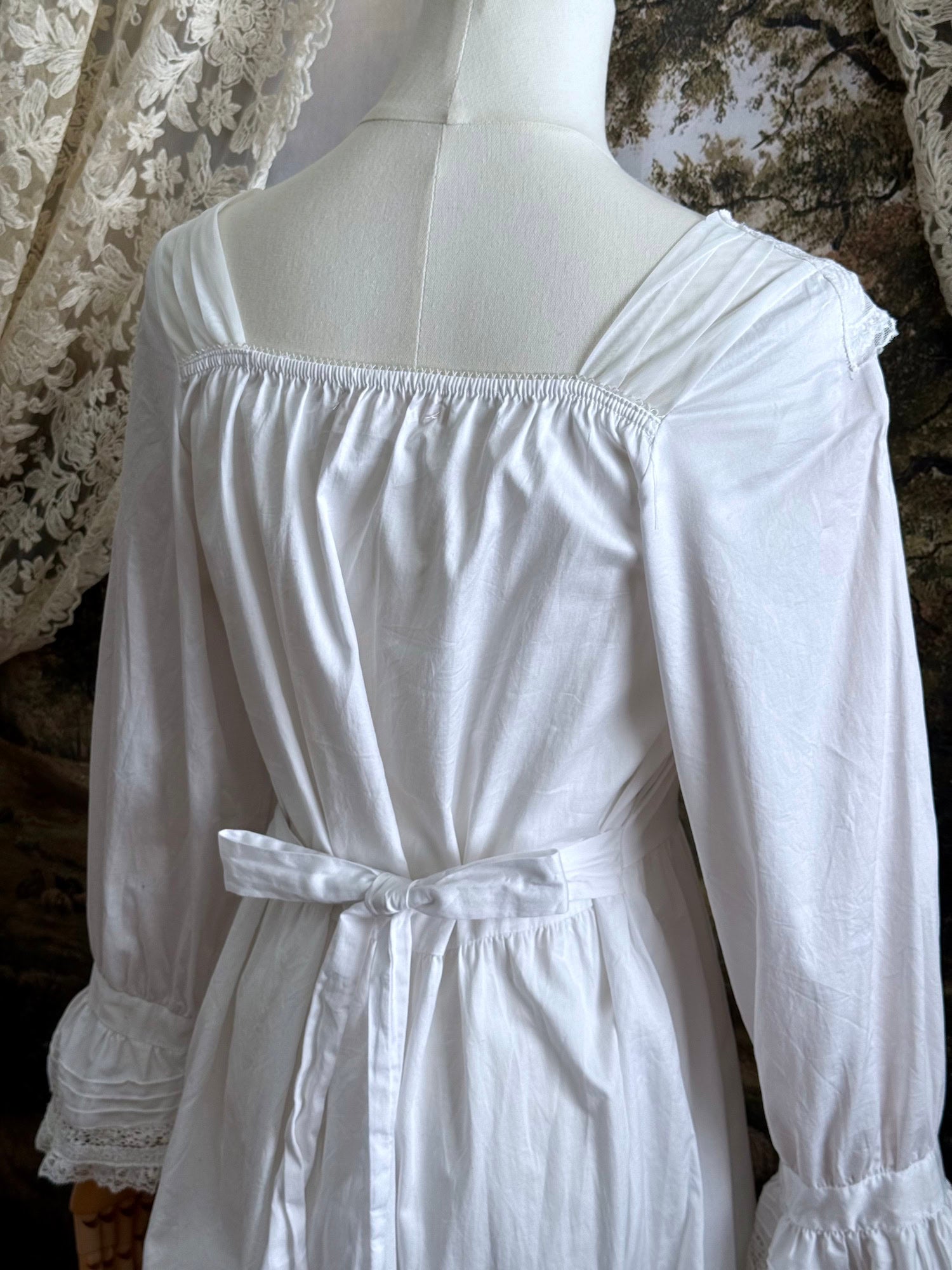 A historically-inspired 1700s style white cotton basque waist night dress with bell sleeves and lace trim pictured on a mannequin in front of an art-history backdrop.