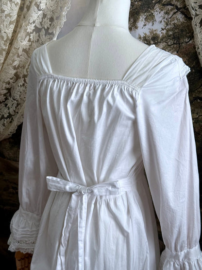A historically-inspired 1700s style white cotton basque waist night dress with bell sleeves and lace trim pictured on a mannequin in front of an art-history backdrop.