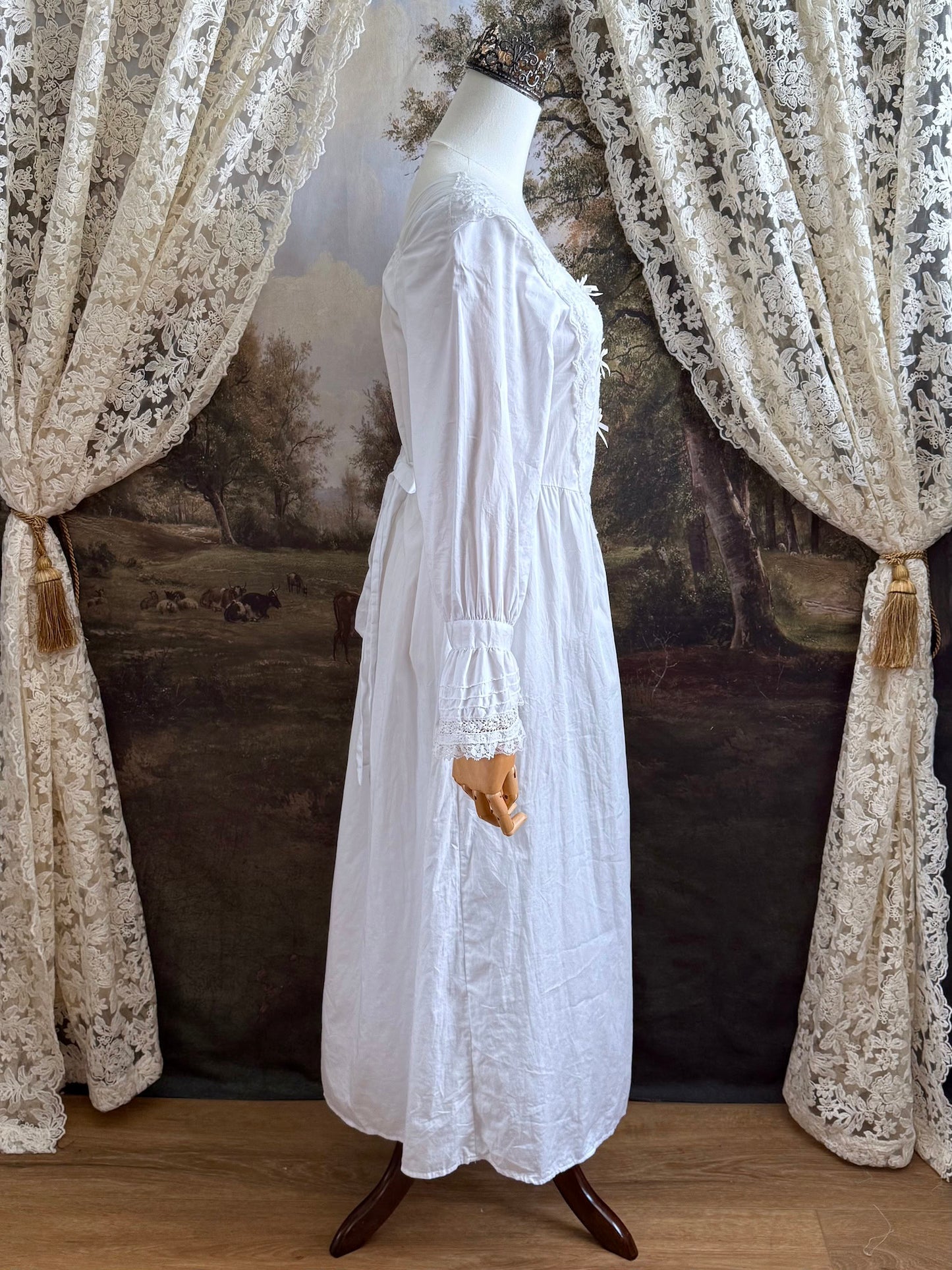 A historically-inspired 1700s style white cotton basque waist night dress with bell sleeves and lace trim pictured on a mannequin in front of an art-history backdrop.