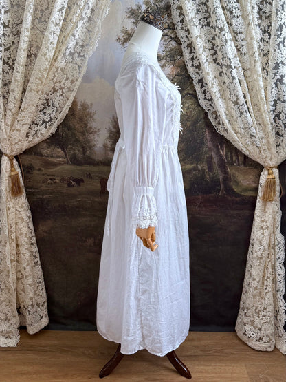 A historically-inspired 1700s style white cotton basque waist night dress with bell sleeves and lace trim pictured on a mannequin in front of an art-history backdrop.