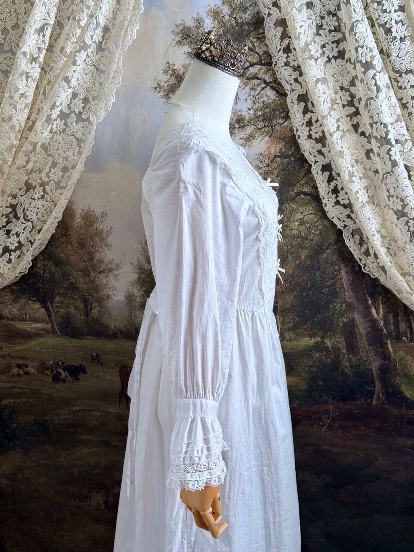 A historically-inspired 1700s style white cotton basque waist night dress with bell sleeves and lace trim pictured on a mannequin in front of an art-history backdrop.