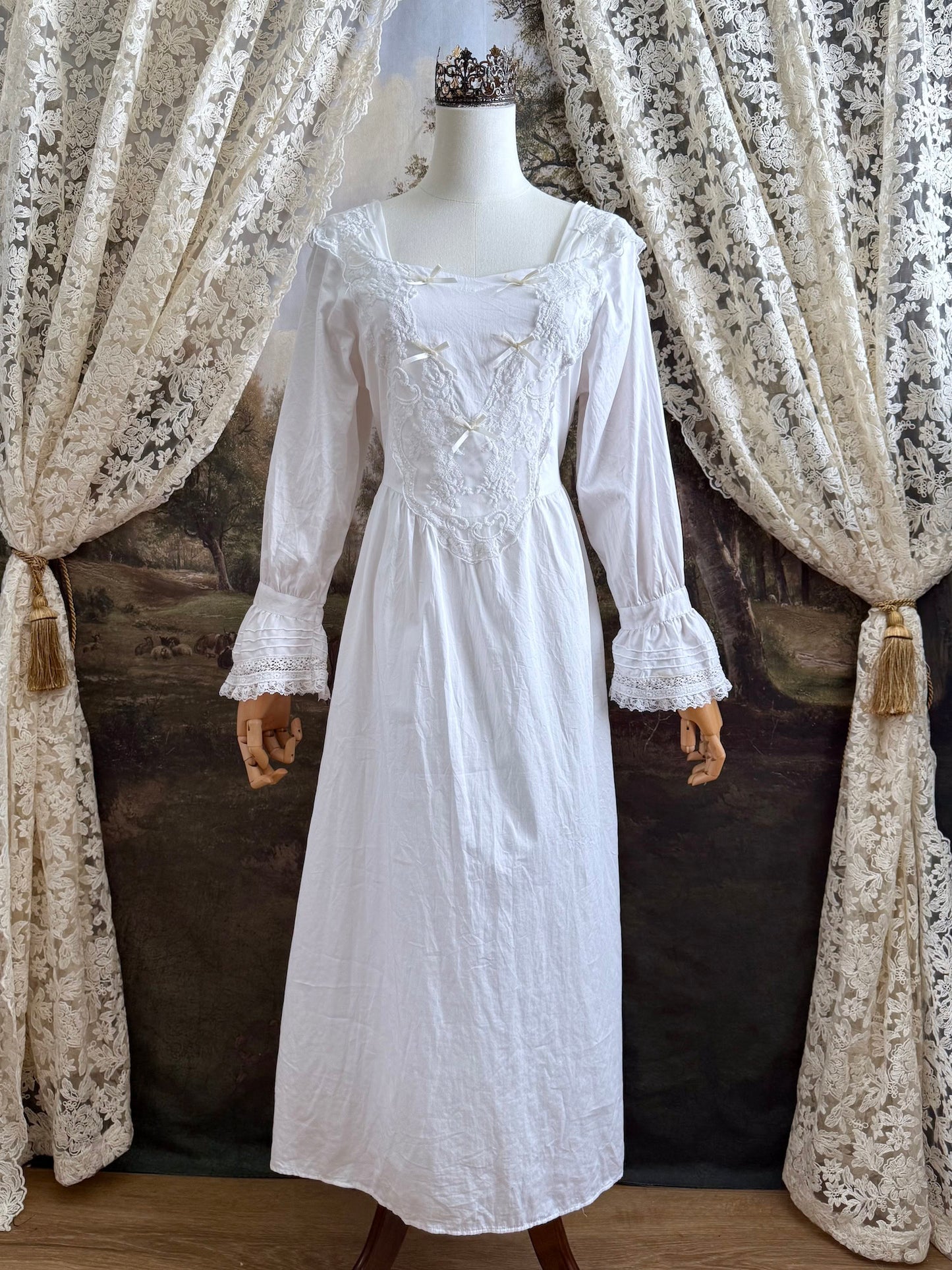 A historically-inspired 1700s style white cotton basque waist night dress with bell sleeves and lace trim pictured on a mannequin in front of an art-history backdrop.