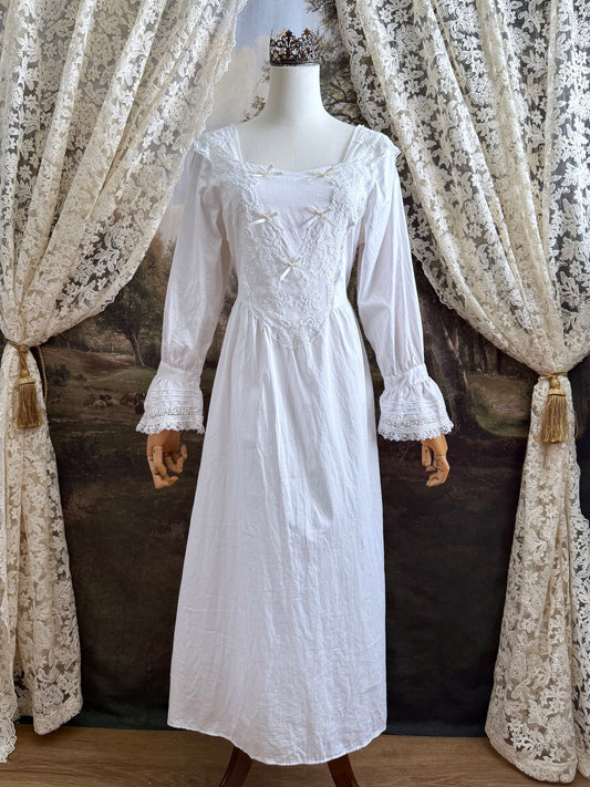 A historically-inspired 1700s style white cotton basque waist night dress with bell sleeves and lace trim pictured on a mannequin in front of an art-history backdrop.