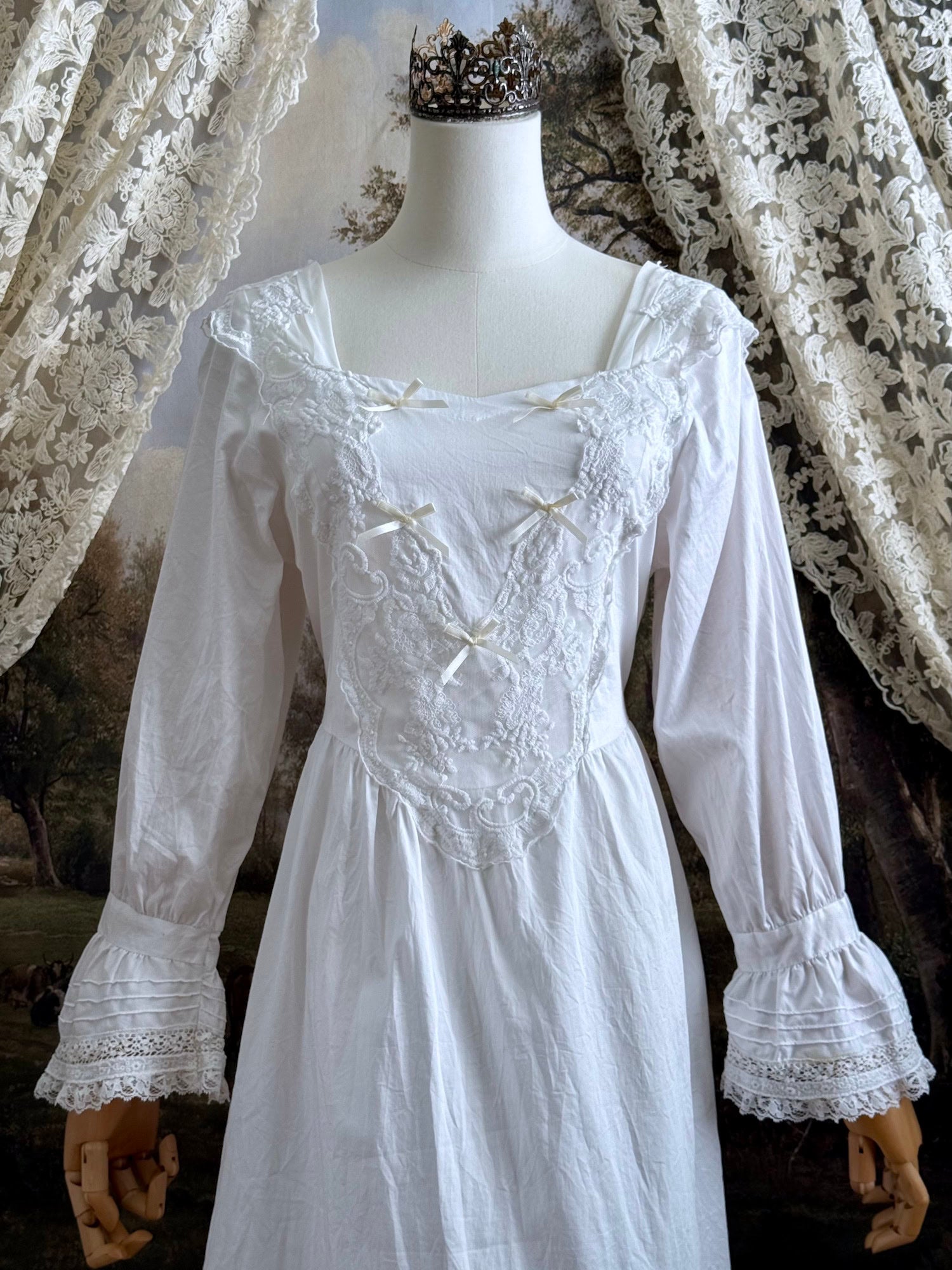 A historically-inspired 1700s style white cotton basque waist night dress with bell sleeves and lace trim pictured on a mannequin in front of an art-history backdrop.