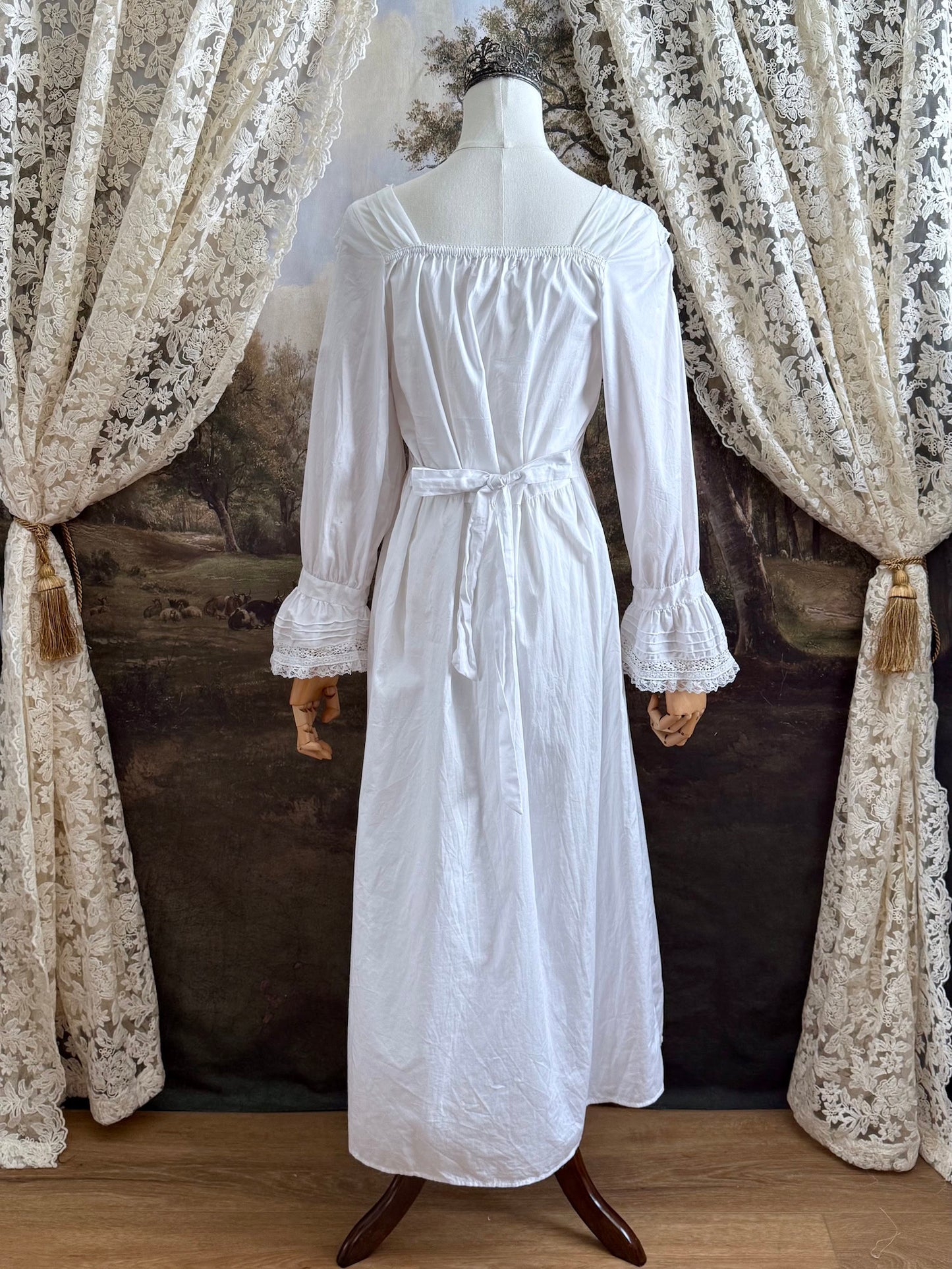 A historically-inspired 1700s style white cotton basque waist night dress with bell sleeves and lace trim pictured on a mannequin in front of an art-history backdrop.