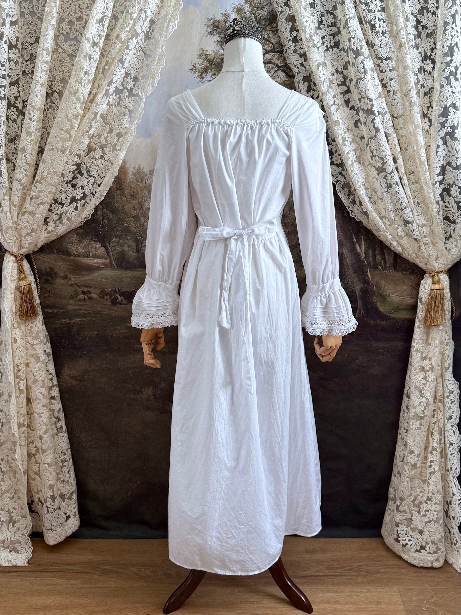 A historically-inspired 1700s style white cotton basque waist night dress with bell sleeves and lace trim pictured on a mannequin in front of an art-history backdrop.