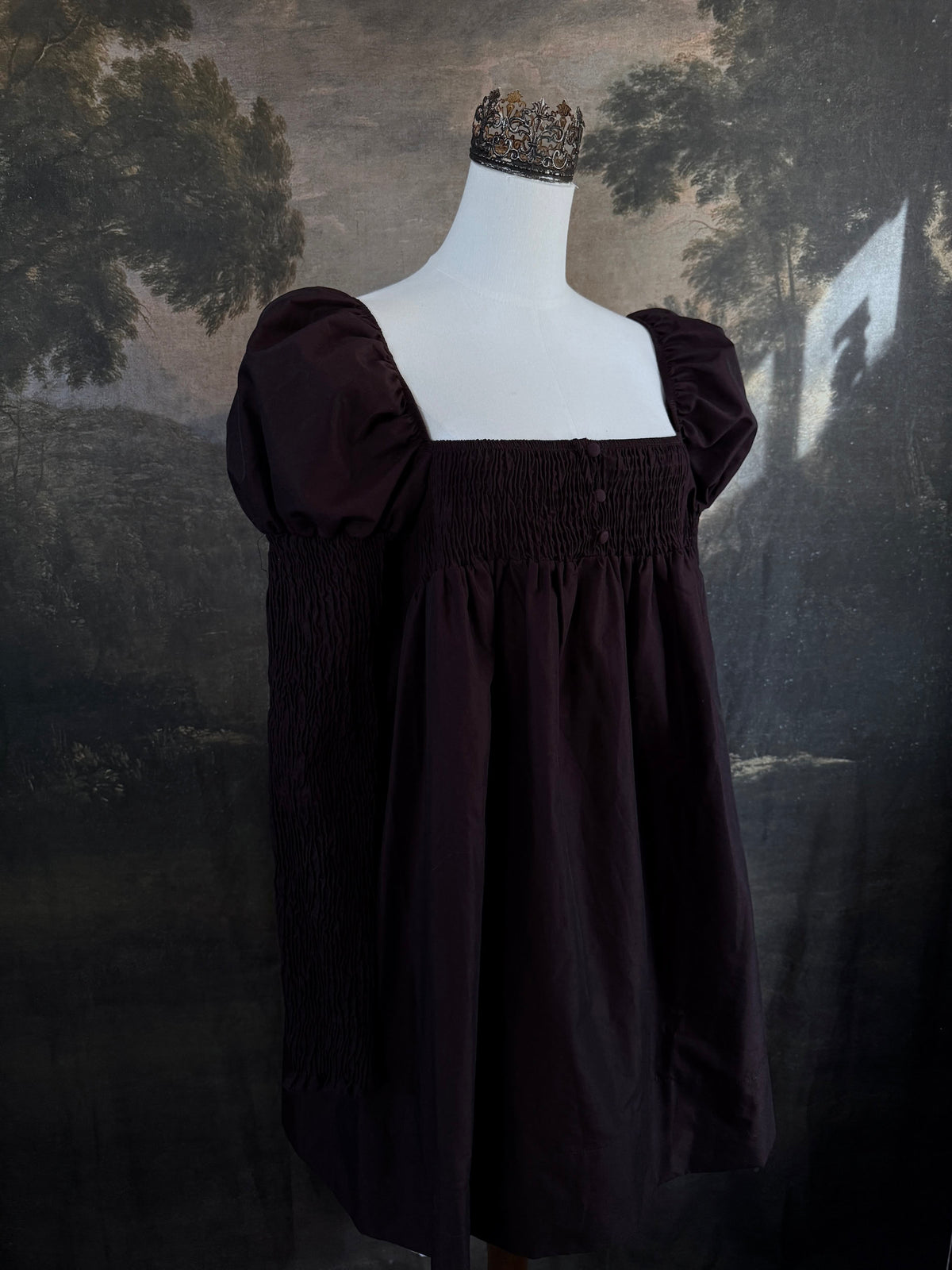 A Historically inspired regency to victorian era puff gigot sleeve mini dress in dark auburn brown is pictured on a mannequin in front of a historic landscape painting backdrop.