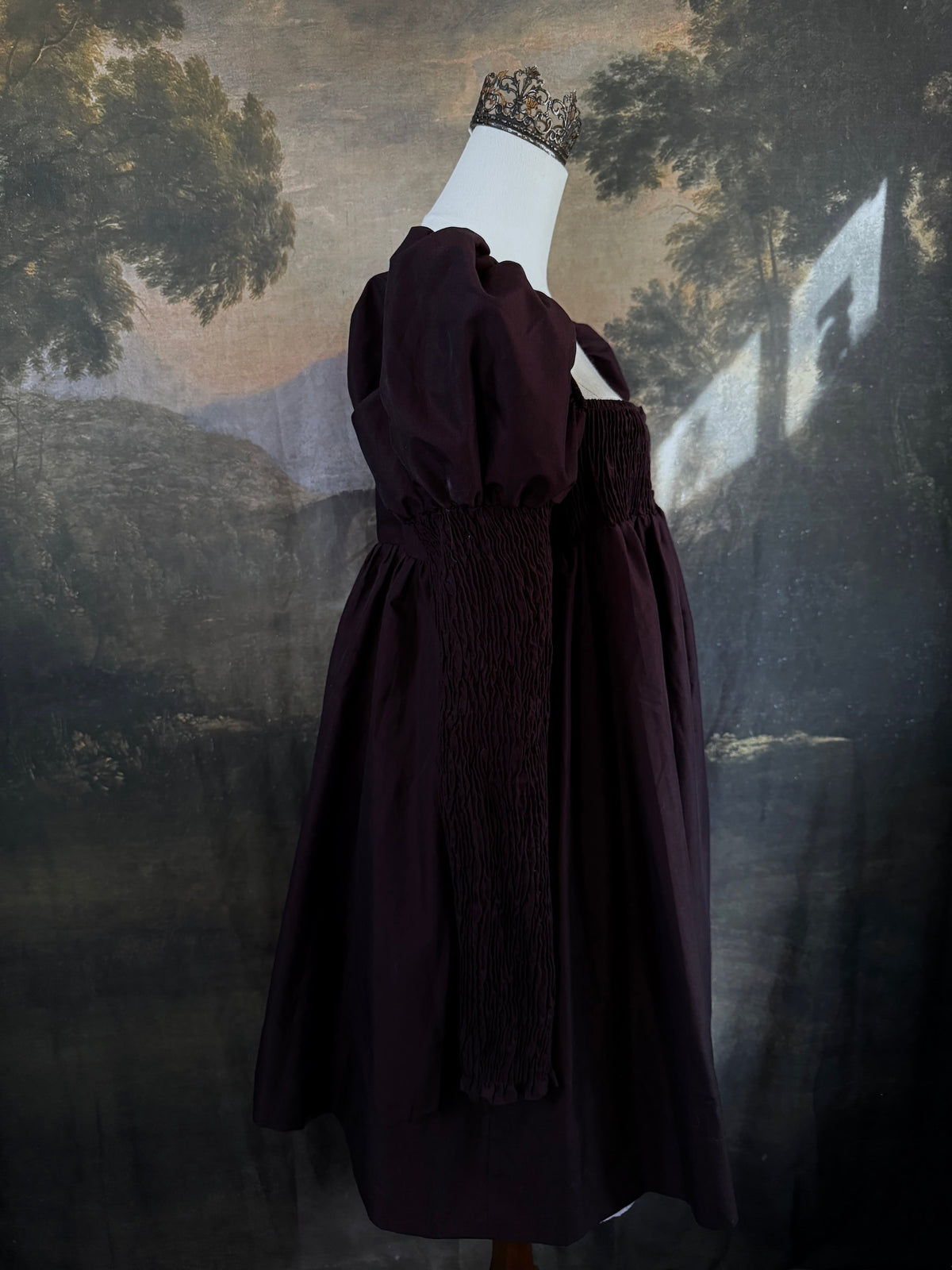 A Historically inspired regency to victorian era puff gigot sleeve mini dress in dark auburn brown is pictured on a mannequin in front of a historic landscape painting backdrop.