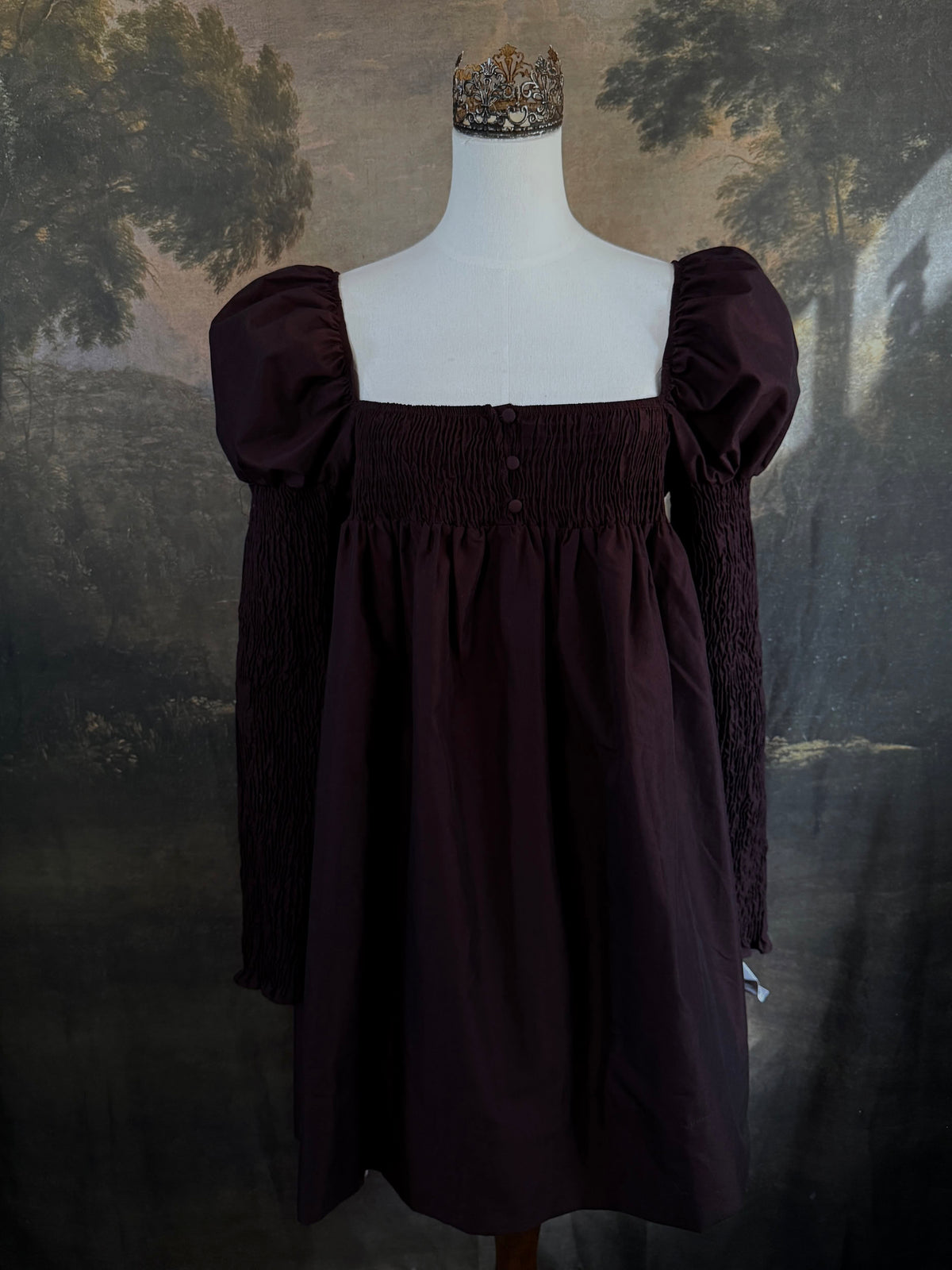A Historically inspired regency to victorian era puff gigot sleeve mini dress in dark auburn brown is pictured on a mannequin in front of a historic landscape painting backdrop.