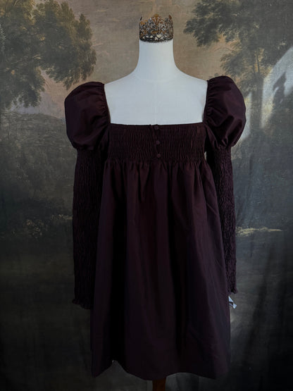 A Historically inspired regency to victorian era puff gigot sleeve mini dress in dark auburn brown is pictured on a mannequin in front of a historic landscape painting backdrop.