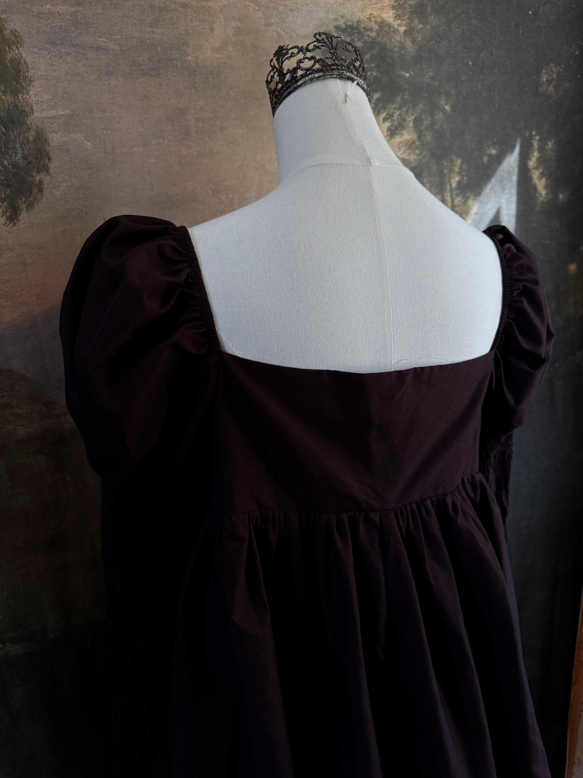 A Historically inspired regency to victorian era puff gigot sleeve mini dress in dark auburn brown is pictured on a mannequin in front of a historic landscape painting backdrop.