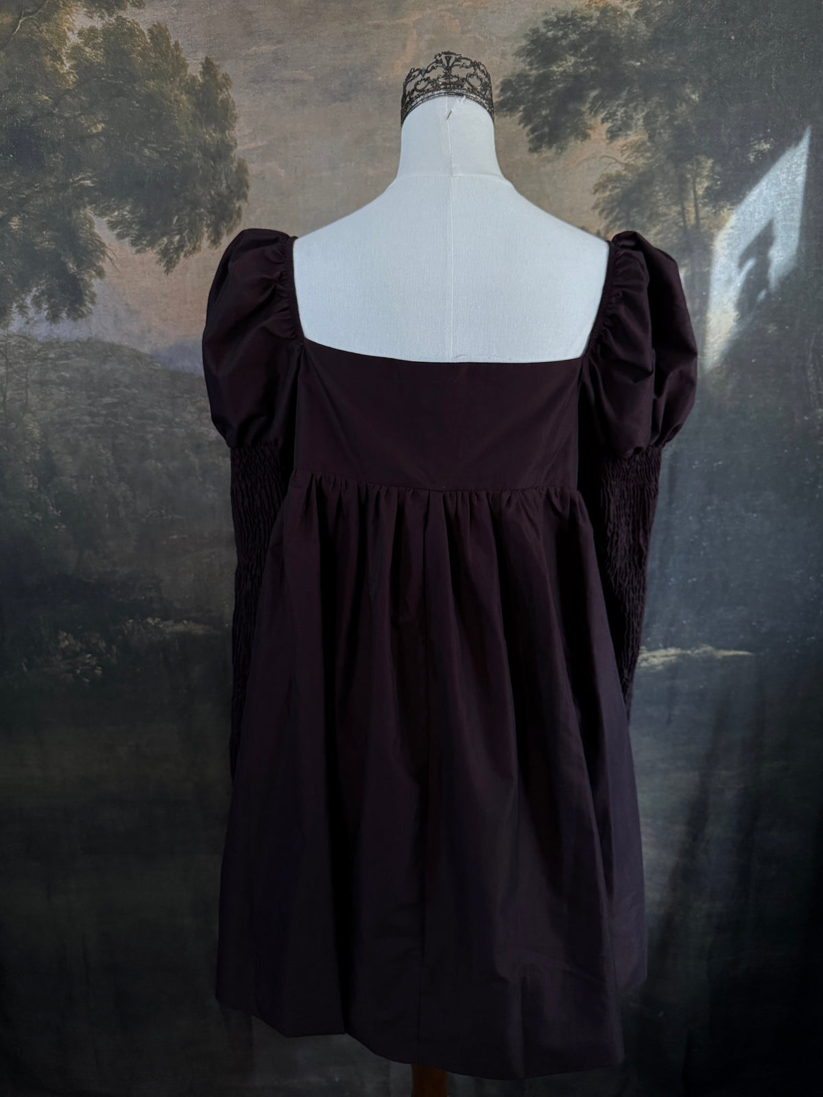 A Historically inspired regency to victorian era puff gigot sleeve mini dress in dark auburn brown is pictured on a mannequin in front of a historic landscape painting backdrop.