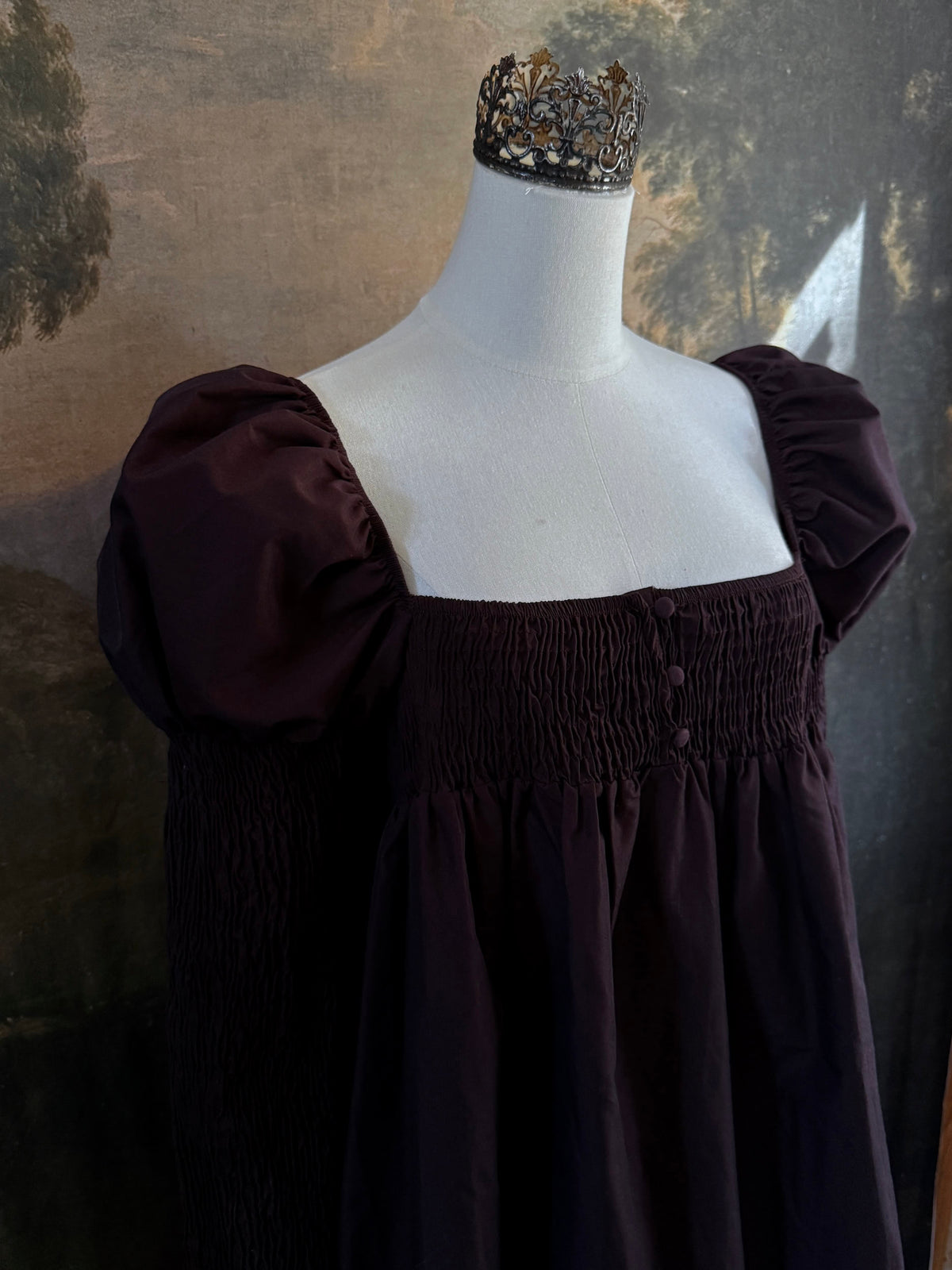 A Historically inspired regency to victorian era puff gigot sleeve mini dress in dark auburn brown is pictured on a mannequin in front of a historic landscape painting backdrop.