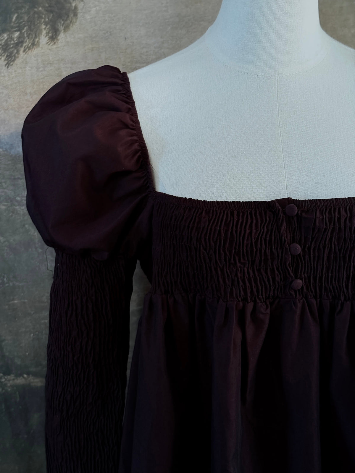 A Historically inspired regency to victorian era puff gigot sleeve mini dress in dark auburn brown is pictured on a mannequin in front of a historic landscape painting backdrop.