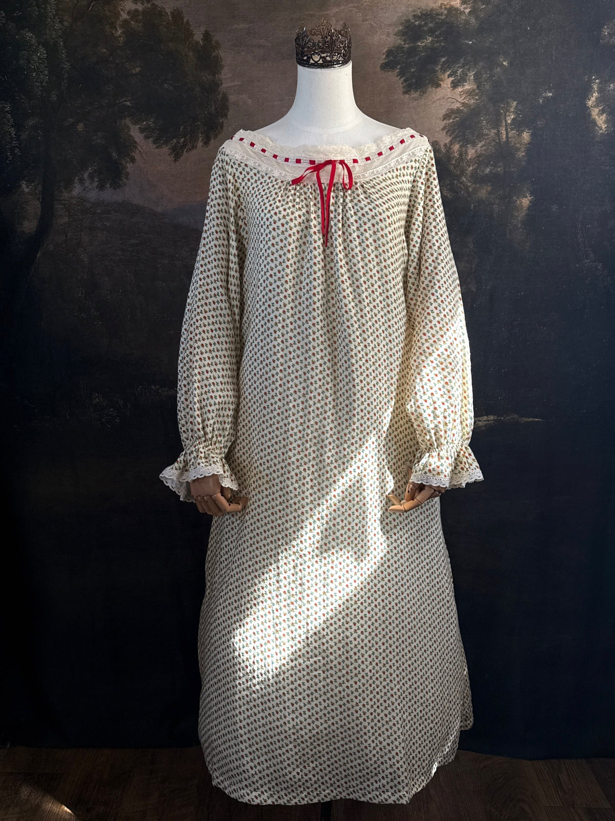 A victorian-inspired historical cream-colored cotton nightgown with a small red and blue fruit pattern, featuring a red ribbon and torchon lace trim, displayed on a mannequin against a dark forest painting backdrop.