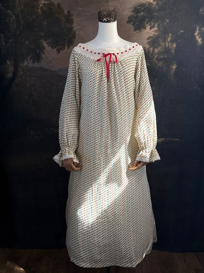 A victorian-inspired historical cream-colored cotton nightgown with a small red and blue fruit pattern, featuring a red ribbon and torchon lace trim, displayed on a mannequin against a dark forest painting backdrop.