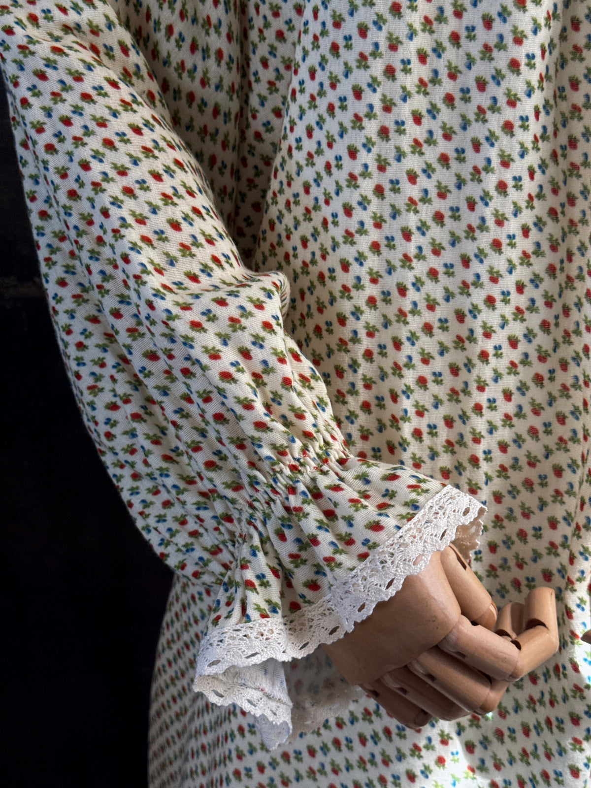 A victorian-inspired historical cream-colored cotton nightgown with a small red and blue fruit pattern, featuring a red ribbon and torchon lace trim, displayed on a mannequin against a dark forest painting backdrop.