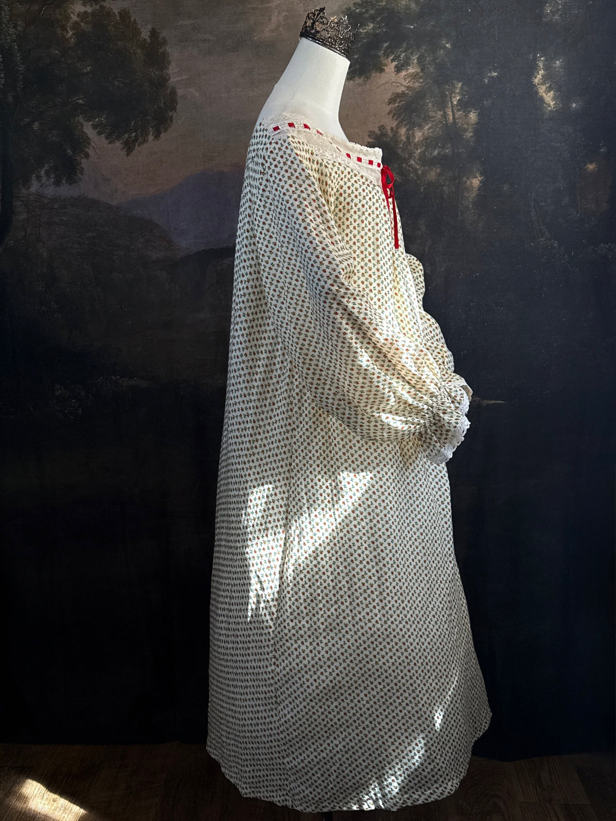 A victorian-inspired historical cream-colored cotton nightgown with a small red and blue fruit pattern, featuring a red ribbon and torchon lace trim, displayed on a mannequin against a dark forest painting backdrop.