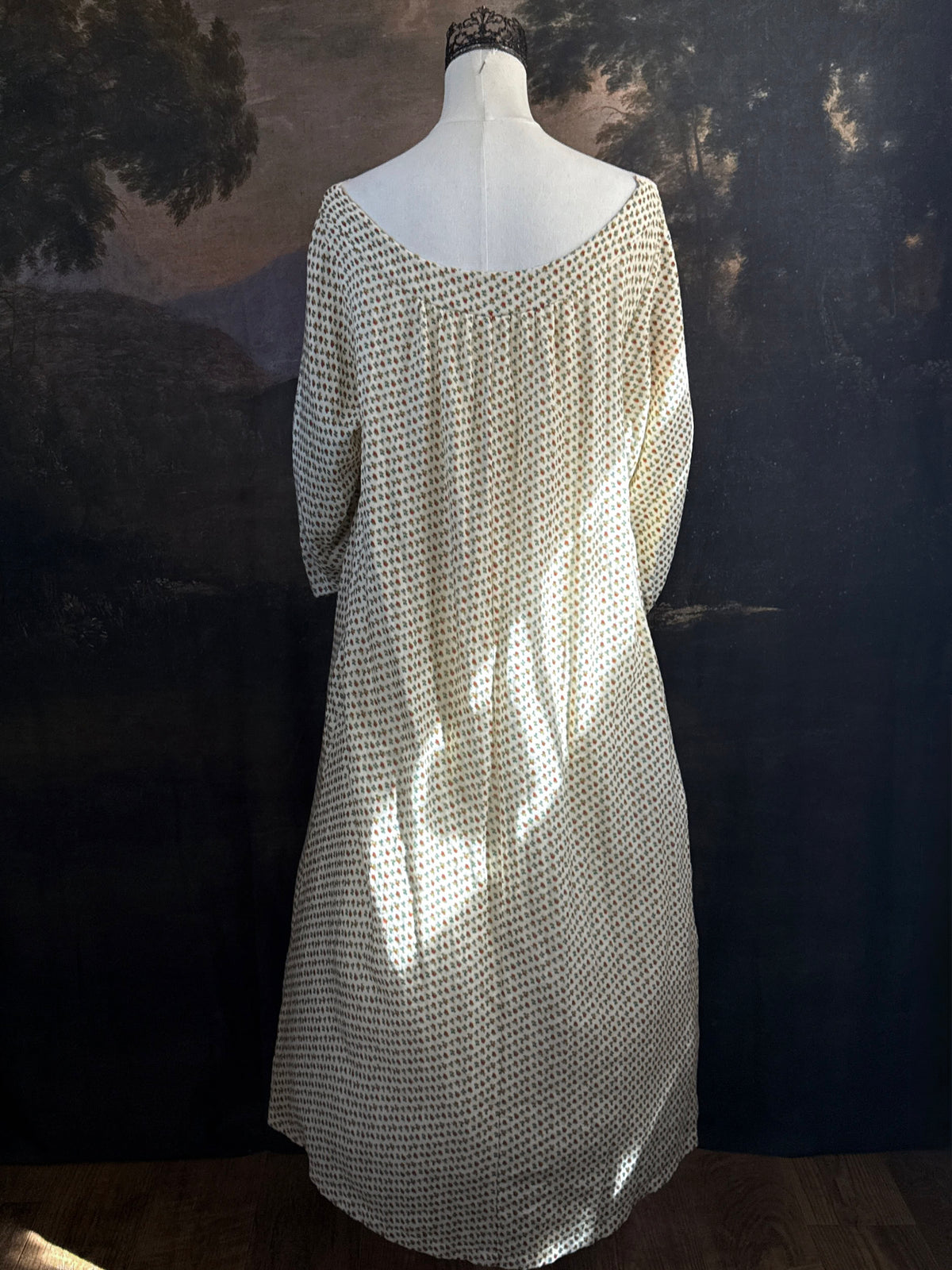 A victorian-inspired historical cream-colored cotton nightgown with a small red and blue fruit pattern, featuring a red ribbon and torchon lace trim, displayed on a mannequin against a dark forest painting backdrop.