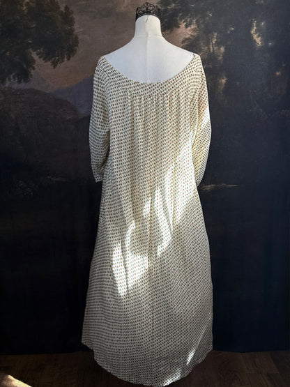 A victorian-inspired historical cream-colored cotton nightgown with a small red and blue fruit pattern, featuring a red ribbon and torchon lace trim, displayed on a mannequin against a dark forest painting backdrop.