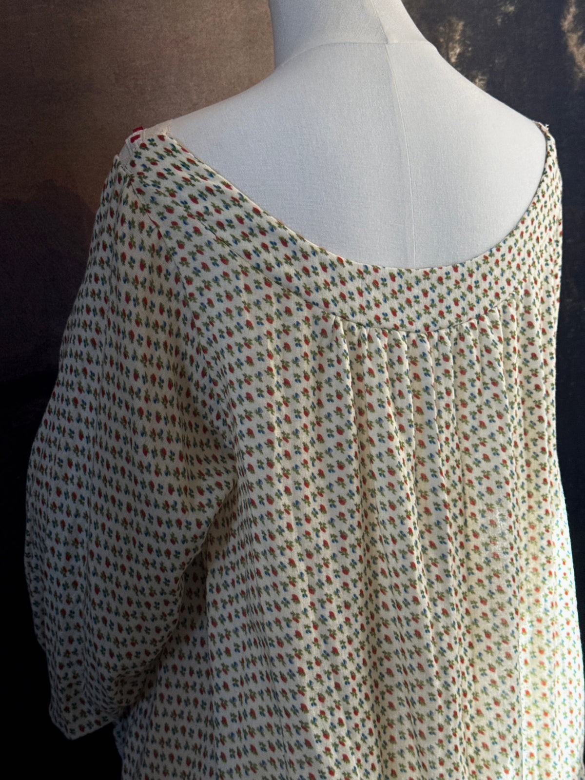 A victorian-inspired historical cream-colored cotton nightgown with a small red and blue fruit pattern, featuring a red ribbon and torchon lace trim, displayed on a mannequin against a dark forest painting backdrop.