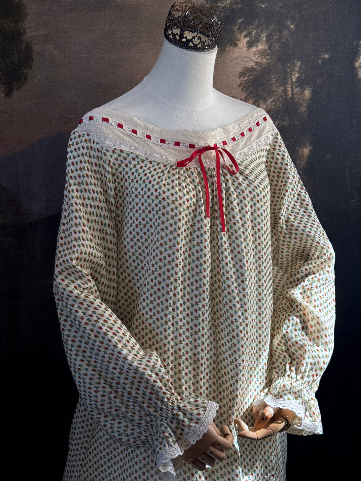A victorian-inspired historical cream-colored cotton nightgown with a small red and blue fruit pattern, featuring a red ribbon and torchon lace trim, displayed on a mannequin against a dark forest painting backdrop.
