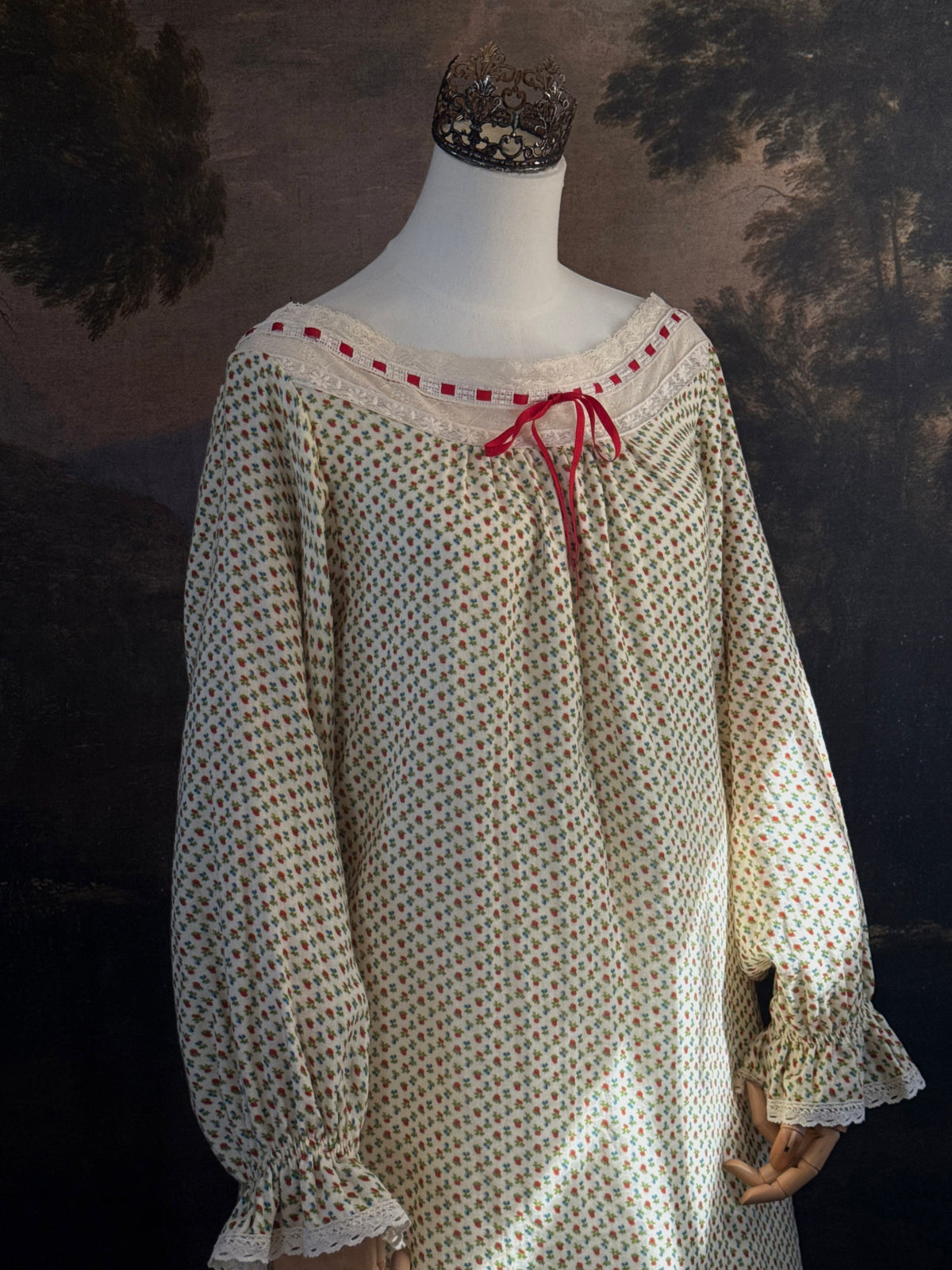 A victorian-inspired historical cream-colored cotton nightgown with a small red and blue fruit pattern, featuring a red ribbon and torchon lace trim, displayed on a mannequin against a dark forest painting backdrop.