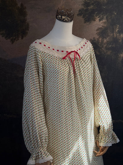 A victorian-inspired historical cream-colored cotton nightgown with a small red and blue fruit pattern, featuring a red ribbon and torchon lace trim, displayed on a mannequin against a dark forest painting backdrop.