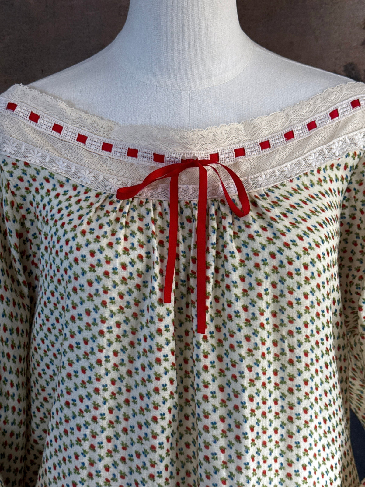 A victorian-inspired historical cream-colored cotton nightgown with a small red and blue fruit pattern, featuring a red ribbon and torchon lace trim, displayed on a mannequin against a dark forest painting backdrop.