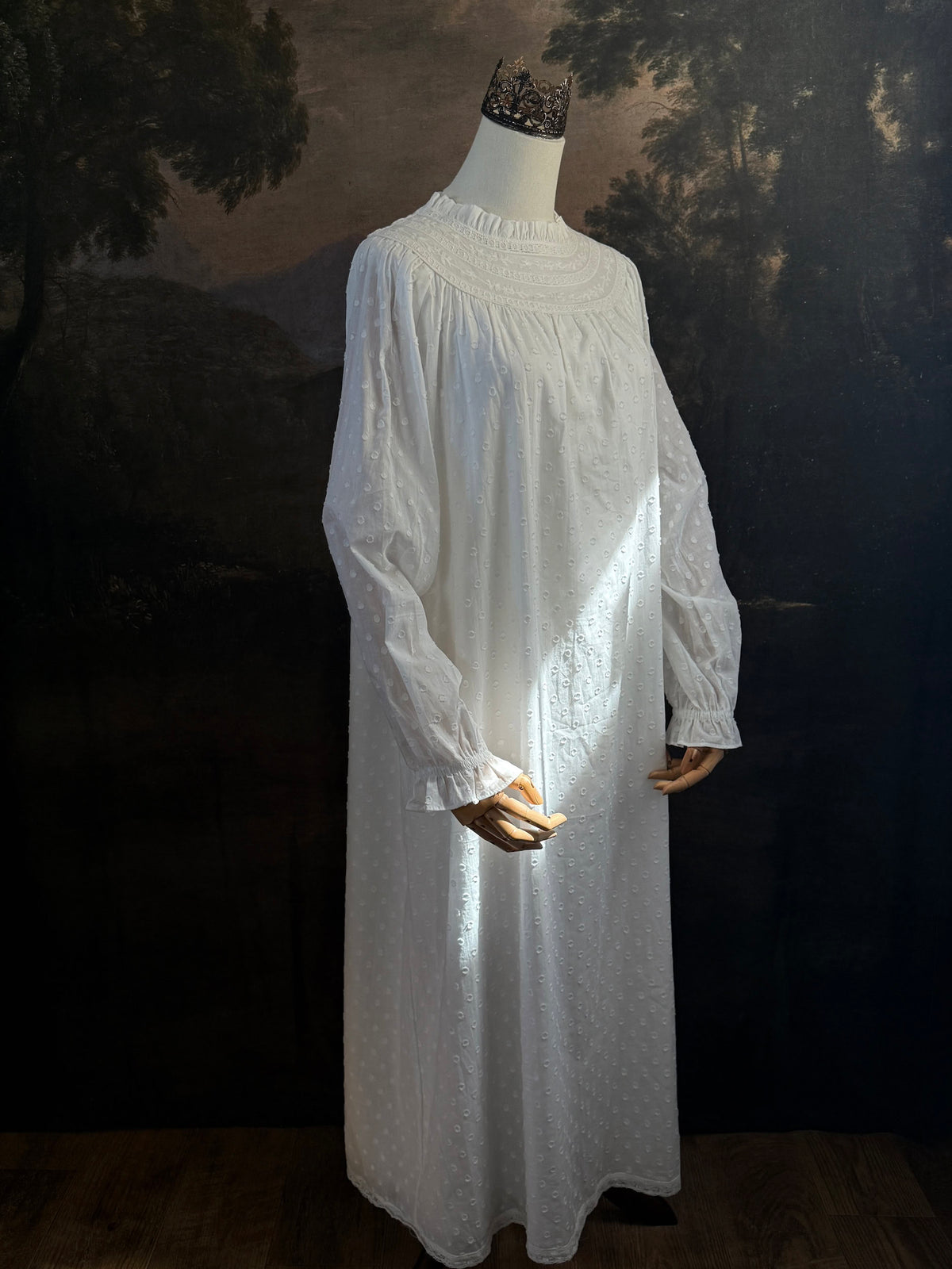 A victorian or edwardian inspired long white cotton nightgown displayed on a mannequin against a dark painted backdrop. The garment has long ruffle end sleeves, a high neckline with lace trim, and a loose, flowing silhouette.