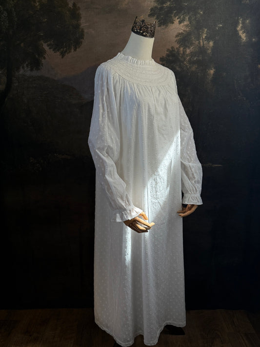 A victorian or edwardian inspired long white cotton nightgown displayed on a mannequin against a dark painted backdrop. The garment has long ruffle end sleeves, a high neckline with lace trim, and a loose, flowing silhouette.