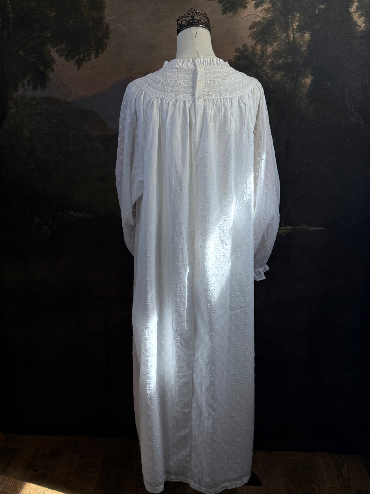 A victorian or edwardian inspired long white cotton nightgown displayed on a mannequin against a dark painted backdrop. The garment has long ruffle end sleeves, a high neckline with lace trim, and a loose, flowing silhouette.