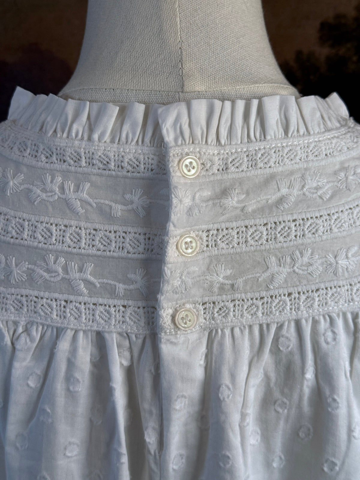 A victorian or edwardian inspired long white cotton nightgown displayed on a mannequin against a dark painted backdrop. The garment has long ruffle end sleeves, a high neckline with lace trim, and a loose, flowing silhouette.