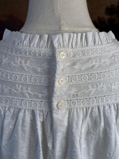 A victorian or edwardian inspired long white cotton nightgown displayed on a mannequin against a dark painted backdrop. The garment has long ruffle end sleeves, a high neckline with lace trim, and a loose, flowing silhouette.