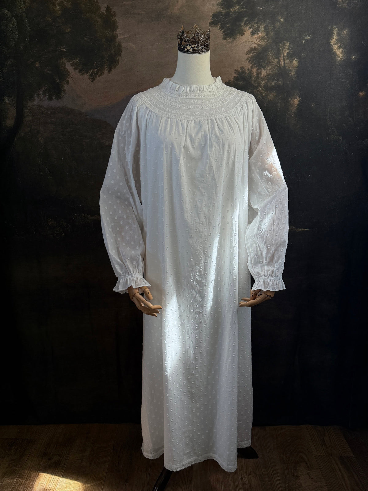 A victorian or edwardian inspired long white cotton nightgown displayed on a mannequin against a dark painted backdrop. The garment has long ruffle end sleeves, a high neckline with lace trim, and a loose, flowing silhouette.