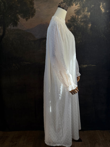 A victorian or edwardian inspired long white cotton nightgown displayed on a mannequin against a dark painted backdrop. The garment has long ruffle end sleeves, a high neckline with lace trim, and a loose, flowing silhouette.
