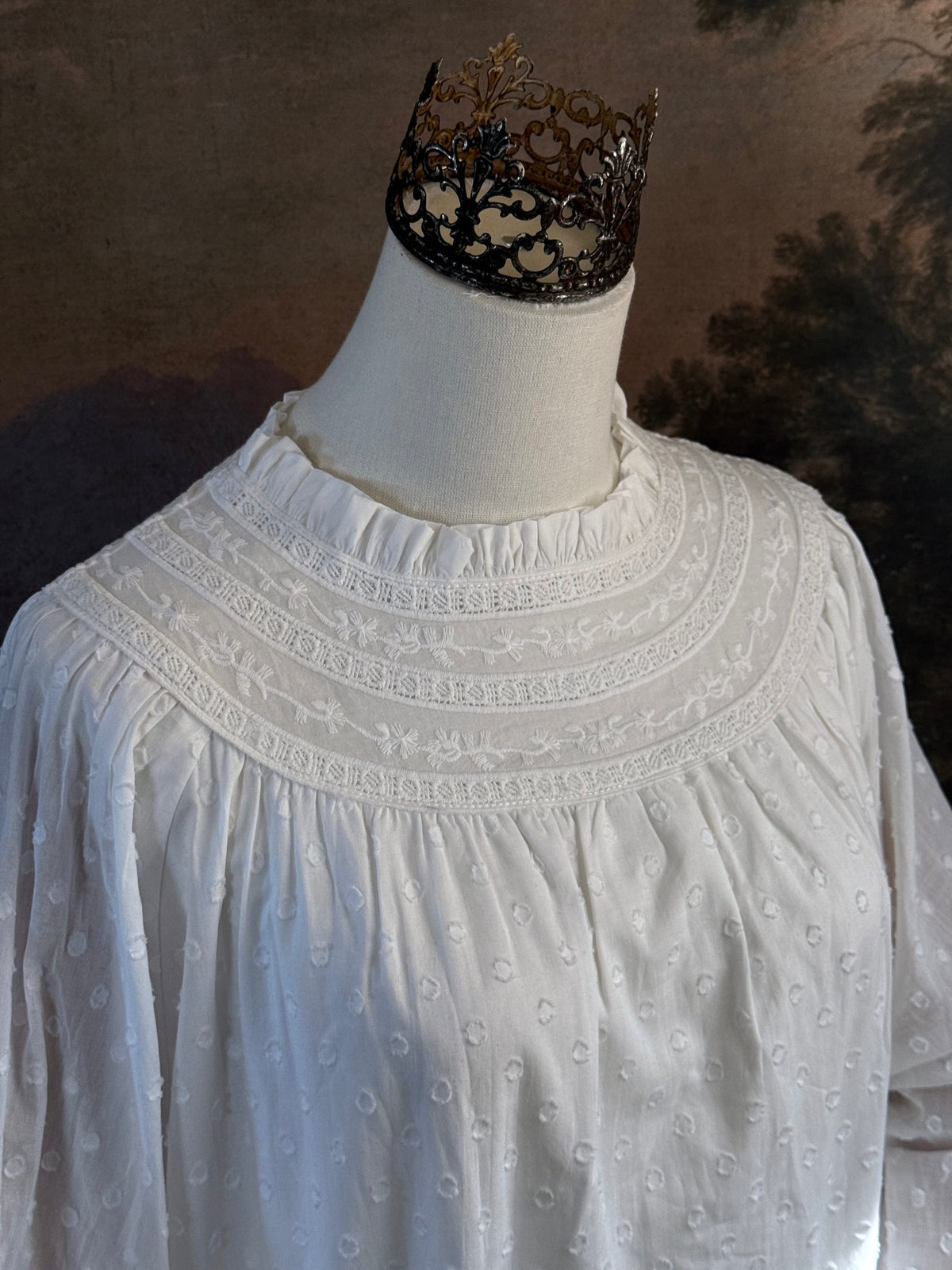 A victorian or edwardian inspired long white cotton nightgown displayed on a mannequin against a dark painted backdrop. The garment has long ruffle end sleeves, a high neckline with lace trim, and a loose, flowing silhouette. Collar detail.