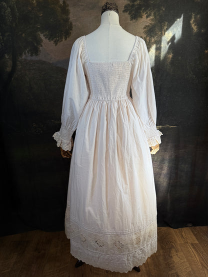 A long ivory/antique-white historical-style chemise dress displayed on a mannequin against a dark painted landscape backdrop. The dress has long sleeves with ruffled cuffs, a smocked bodice, a lace-trimmed neckline with front corset lacing, and lace inset details along the hem.