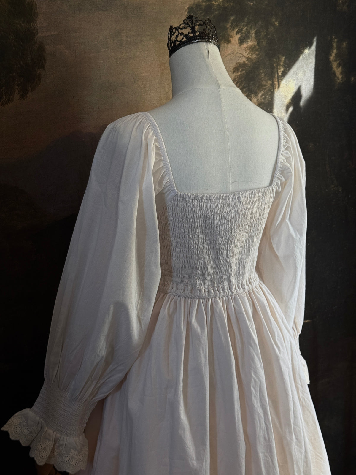 A long ivory/antique-white historical-style chemise dress displayed on a mannequin against a dark painted landscape backdrop. The dress has long sleeves with ruffled cuffs, a smocked bodice, a lace-trimmed neckline with front corset lacing, and lace inset details along the hem.