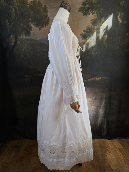 A long ivory/antique-white historical-style chemise dress displayed on a mannequin against a dark painted landscape backdrop. The dress has long sleeves with ruffled cuffs, a smocked bodice, a lace-trimmed neckline with front corset lacing, and lace inset details along the hem.