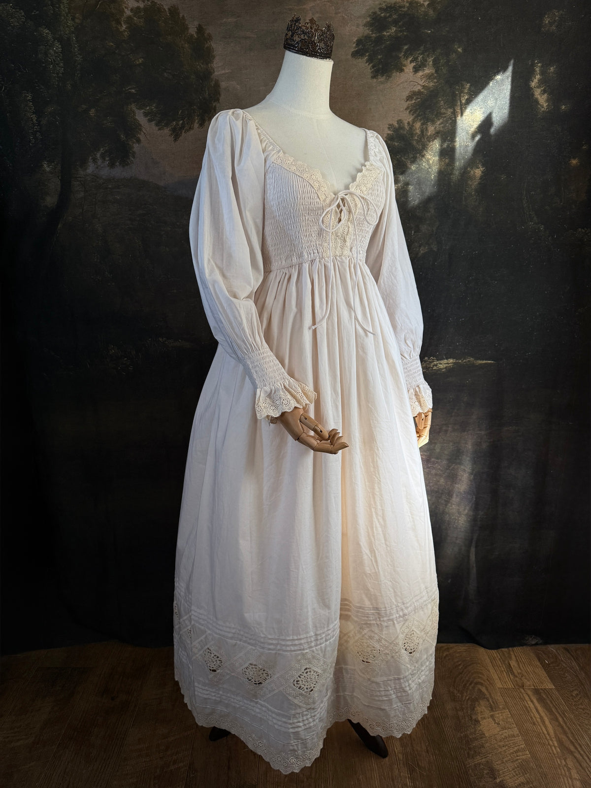 A long ivory/antique-white historical-style chemise dress displayed on a mannequin against a dark painted landscape backdrop. The dress has long sleeves with ruffled cuffs, a smocked bodice, a lace-trimmed neckline with front corset lacing, and lace inset details along the hem.