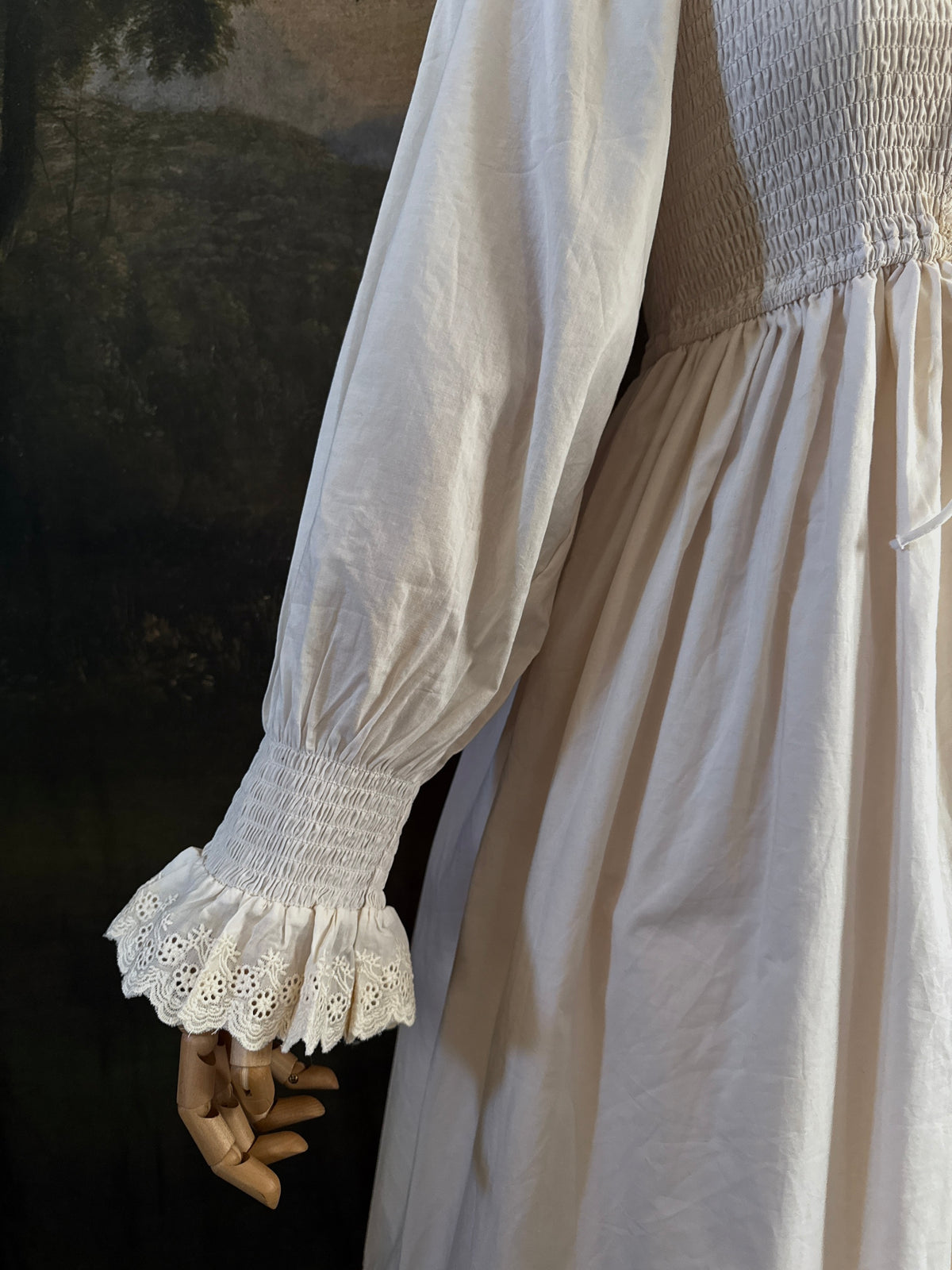 A long ivory/antique-white historical-style chemise dress displayed on a mannequin against a dark painted landscape backdrop. The dress has long sleeves with ruffled cuffs, a smocked bodice, a lace-trimmed neckline with front corset lacing, and lace inset details along the hem.
