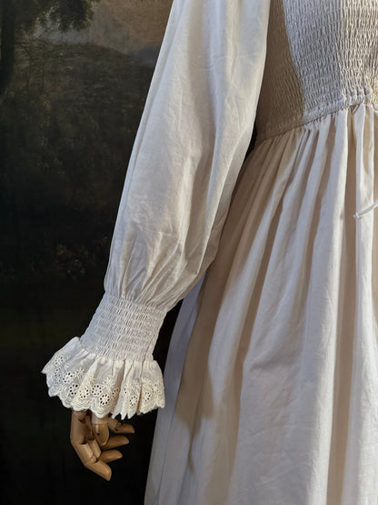 A long ivory/antique-white historical-style chemise dress displayed on a mannequin against a dark painted landscape backdrop. The dress has long sleeves with ruffled cuffs, a smocked bodice, a lace-trimmed neckline with front corset lacing, and lace inset details along the hem.