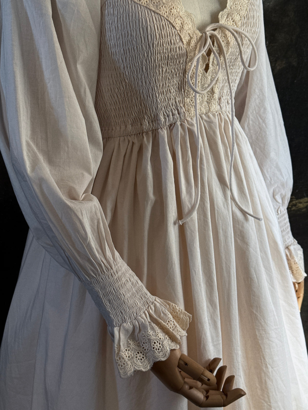 A long ivory/antique-white historical-style chemise dress displayed on a mannequin against a dark painted landscape backdrop. The dress has long sleeves with ruffled cuffs, a smocked bodice, a lace-trimmed neckline with front corset lacing, and lace inset details along the hem. Close up on sleeves.