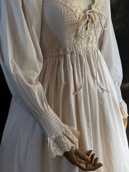 A long ivory/antique-white historical-style chemise dress displayed on a mannequin against a dark painted landscape backdrop. The dress has long sleeves with ruffled cuffs, a smocked bodice, a lace-trimmed neckline with front corset lacing, and lace inset details along the hem. Close up on sleeves.