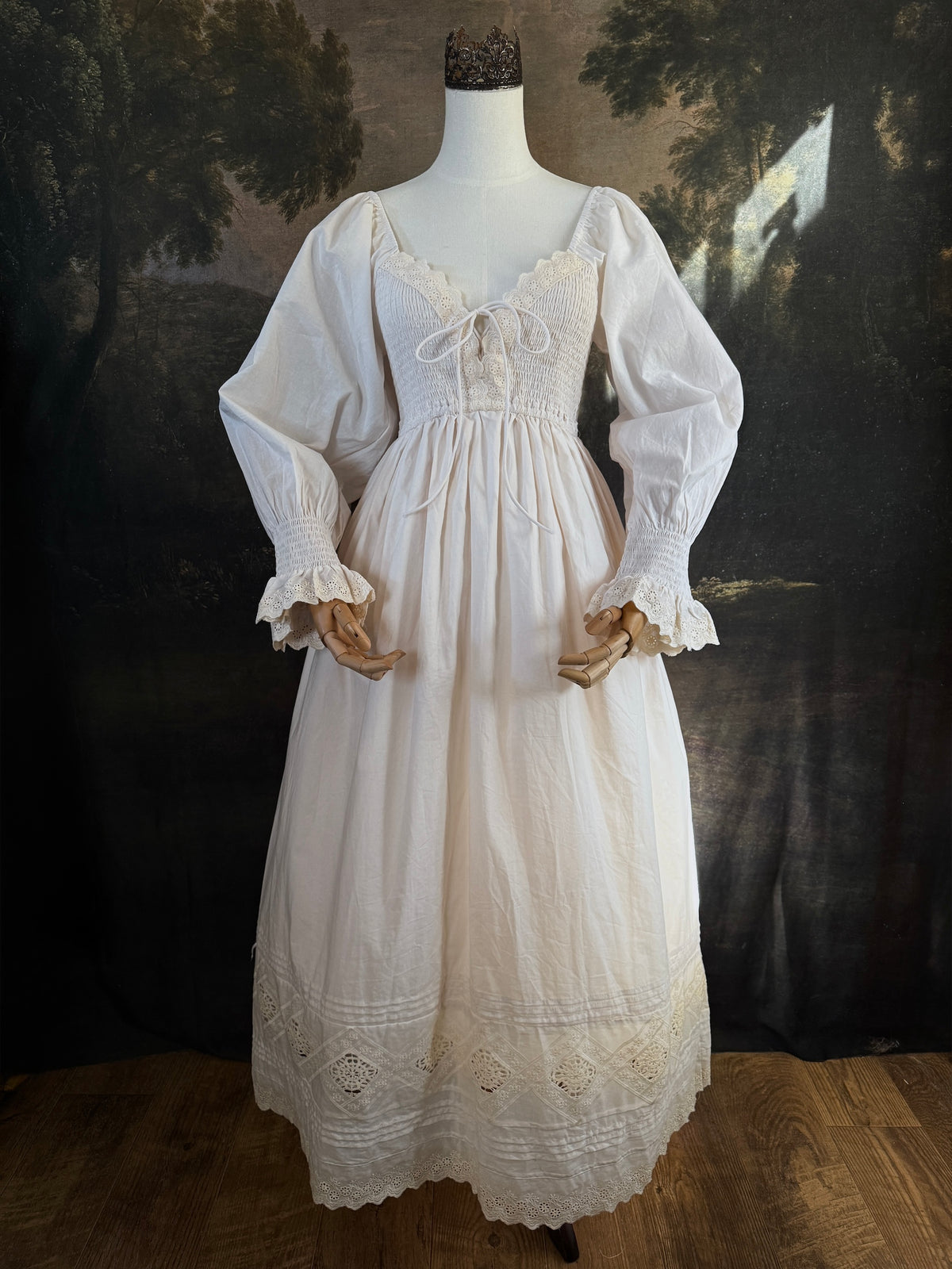 A long ivory/antique-white historical-style chemise dress displayed on a mannequin against a dark painted landscape backdrop. The dress has long sleeves with ruffled cuffs, a smocked bodice, a lace-trimmed neckline with front corset lacing, and lace inset details along the hem.